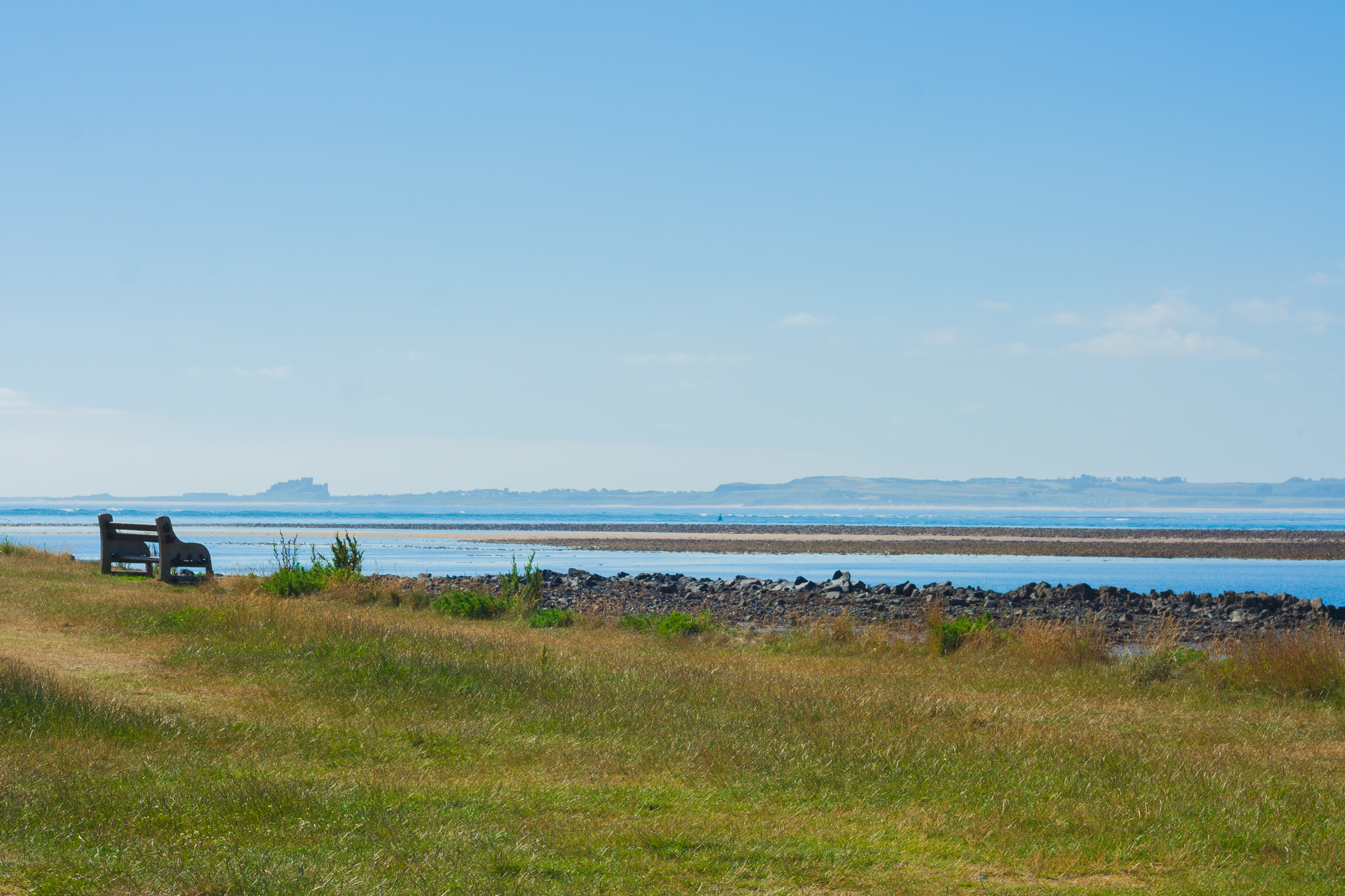 A bench on a grassy bank overlooking a sandy bank and the sea on a sunny day