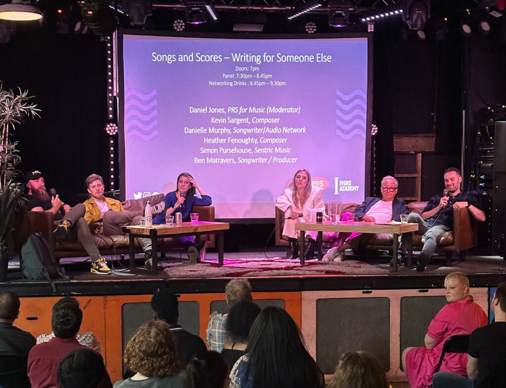 Songs And Scores Writing For Someone Else stage with six seated composers and producers on a panel. One speaks into a microphone whilst the rest look into the audience.